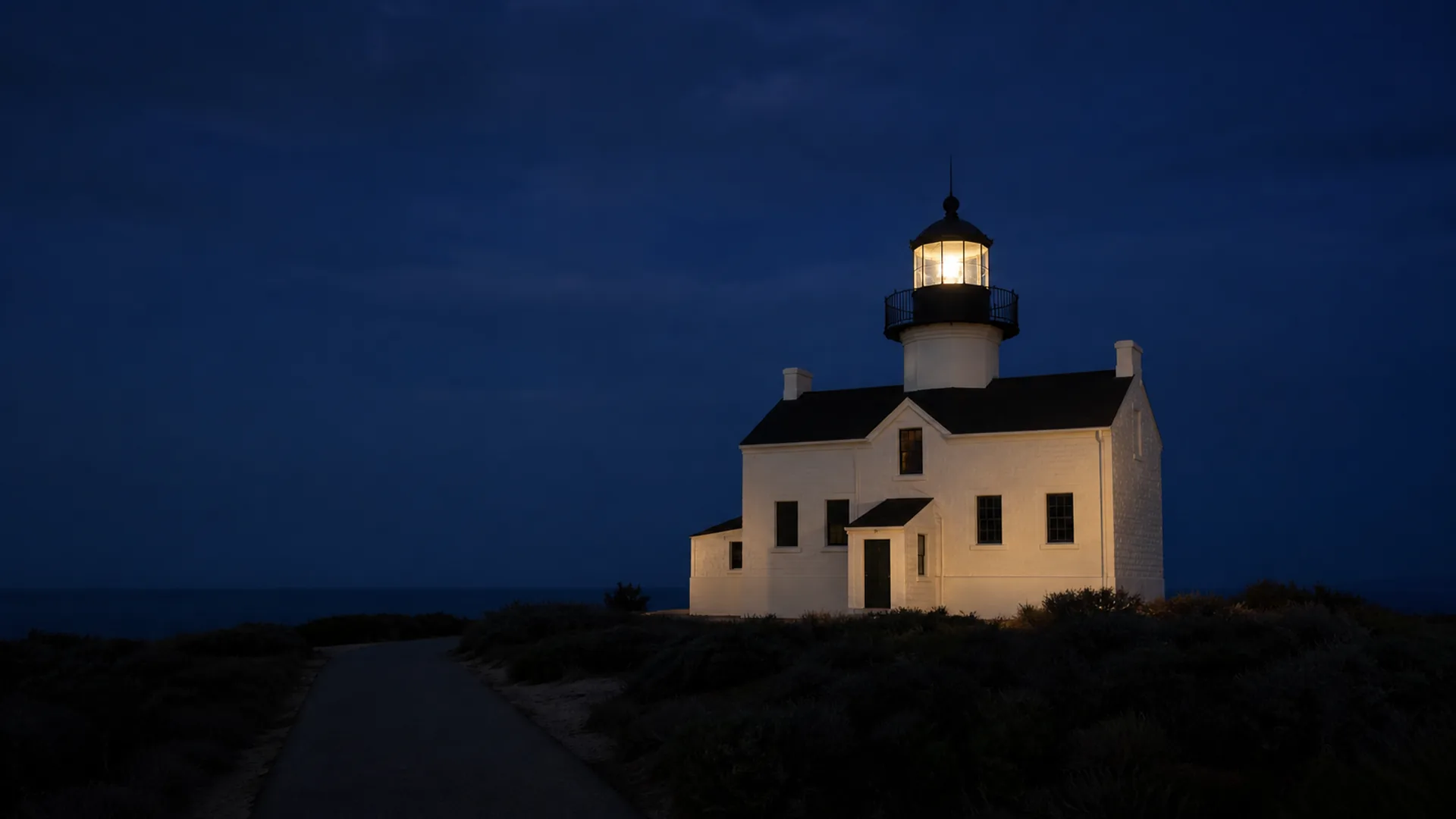 Old Point Loma Lighthouse stands above dark coastal scrub at blue hour.