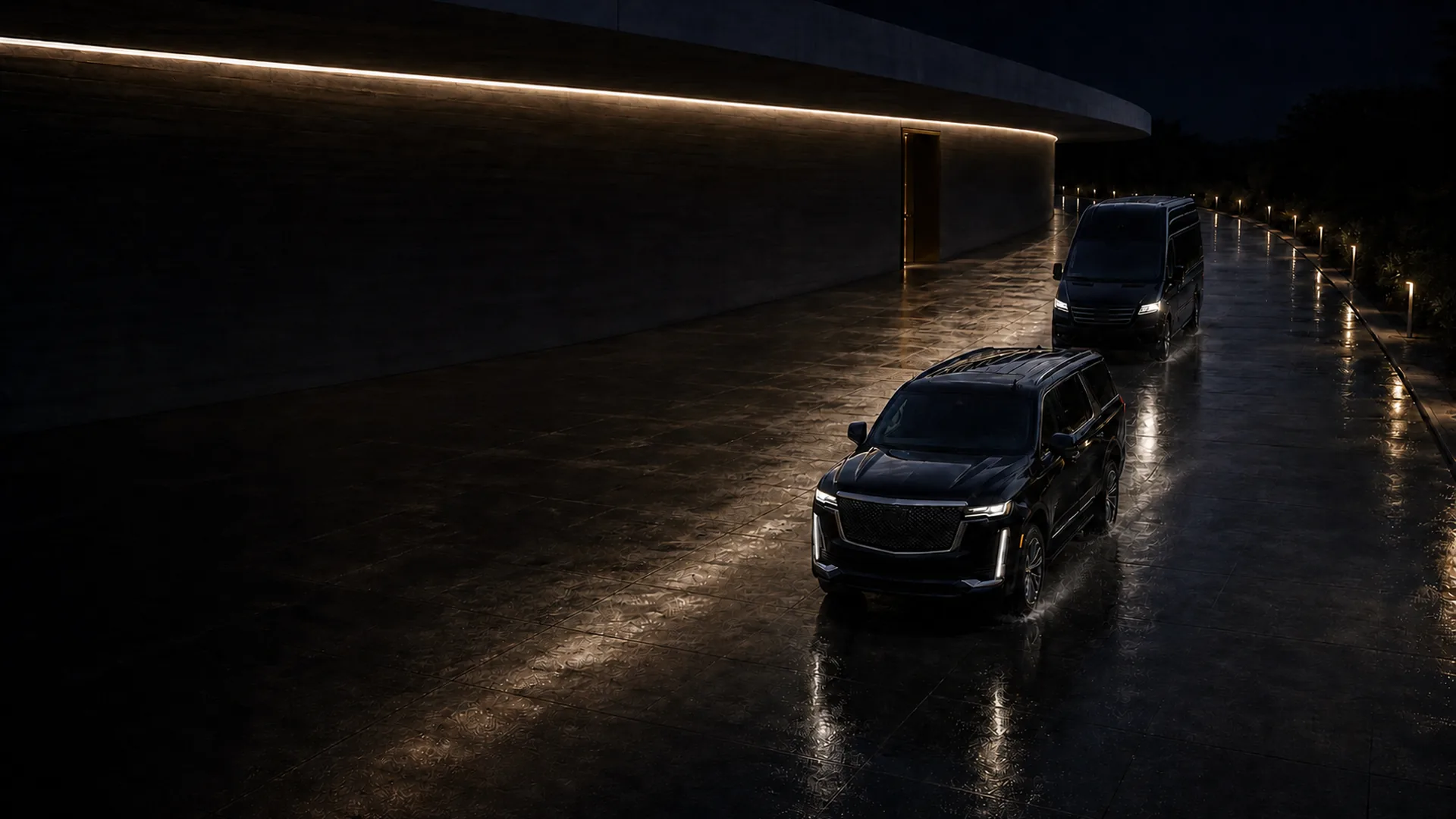 A black Cadillac Escalade followed by a black Mercedes-Benz Sprinter van driving along a wet courtyard at night beside a long lit building.